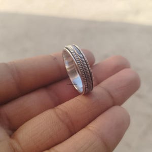 Puede incluir: Un anillo de metal plateado que presenta una banda central con una textura de cuerda trenzada, flanqueada por dos bordes lisos y elevados. El anillo tiene un acabado pulido y un ancho constante en todo su diseño circular detallado.