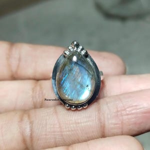 May include: A silver ring featuring a pear-shaped, iridescent labradorite gemstone. The stone displays blue and gold flashes. The ring has a decorative bezel and small beaded accents. The ring is held in a hand.