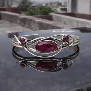 May include: Silver cuff bracelet with three oval-cut, deep red gemstones. The bracelet features an open design with swirling silver accents and is reflected on a dark, glossy surface.