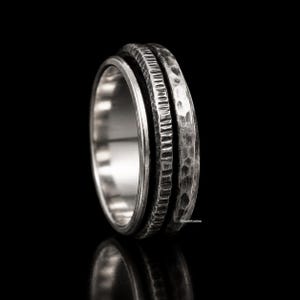 Puede incluir: Un anillo de metal en tono plateado con un diseño de bandas múltiples. La superficie exterior presenta dos texturas distintas: una sección con un patrón acanalado vertical y una sección adyacente con un acabado martillado. La banda interior es lisa.