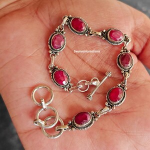 Può includere: Un braccialetto d'argento con pietre preziose rosse ovali e vibranti. Il braccialetto ha un design ornato con una chiusura a levetta e tre anelli collegati. Il testo "Swaroskicreations" è visibile.
