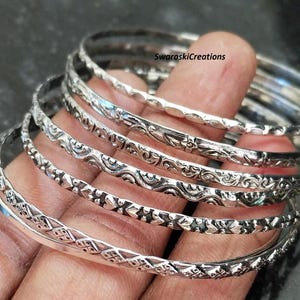 May include: Set of seven silver bangles with intricate floral and geometric designs. The bangles are stacked on a person's arm.