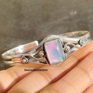 May include: A silver bracelet with an iridescent opal-like stone set in a delicate, ornate design. The bracelet is open and adjustable.