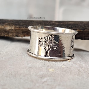 May include: A wide, silver-colored ring with a tree design. The ring has a textured band with a detailed tree carving. The ring is displayed on a light gray surface.