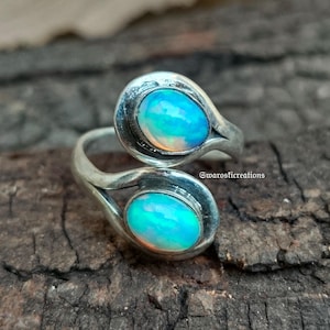 May include: A silver-toned ring with two oval-shaped, iridescent opal stones. The opals display a mix of blue, green, and white hues. The ring's design features a swirling, open-ended band that cradles each stone. The ring is set against a dark, textured background.