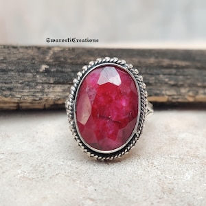May include: A silver ring featuring a large, oval-cut ruby gemstone. The ruby is a deep red color and is surrounded by a decorative silver setting with a rope-like border. The ring is displayed on a neutral surface.