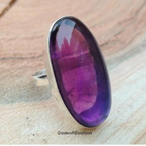 Peut inclure: Une bague en argent ornée d'une grande pierre d'améthyste ovale. L'améthyste présente un dégradé de couleurs violettes, allant du violet foncé aux tons plus clairs. L'anneau de la bague est en argent poli.