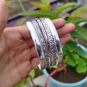 May include: Four silver bangles with detailed floral and geometric designs. The bangles are shown against green foliage, highlighting their polished, reflective surfaces. Each bangle is approximately 1.3 cm wide.