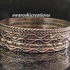 Puede incluir: Un conjunto de cinco pulseras de plata con intrincados diseños en relieve. Las pulseras están apiladas una encima de la otra, creando un aspecto de capas.