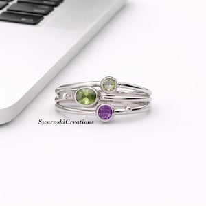 Può includere: Un anello d'argento con tre pietre preziose: una pietra verde ovale, una pietra verde rotonda più piccola e una pietra viola rotonda. L'anello ha più fasce. Il testo "Swaroski Creations" è visibile.