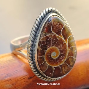 Può includere: Un anello d'argento con una pietra preziosa di ammonite marrone e bianca incastonata in una lunetta a forma di lacrima. L'anello ha un design a corda attorno alla lunetta.
