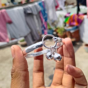 Pode incluir: Uma pulseira prateada com duas pequenas campainhas redondas penduradas em um anel. A pulseira é segurada em uma mão, com roupas penduradas em um varal ao fundo.