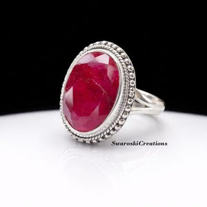 Puede incluir: Un anillo de plata con una gran gema de rubí de forma ovalada. El rubí es de color rojo intenso y está rodeado por un borde de cuentas. El anillo tiene un diseño de caña dividida. El texto "Swaroski Creations" es visible.