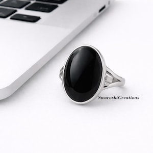 May include: A silver ring with a large, oval, black onyx stone. The ring has a split band design. The ring is on a white surface next to a laptop. The text "SwaroskiCreations" is visible.