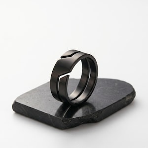 May include: A matte black ring featuring a distinct angular cut-out design that creates a split, layered appearance. The band has a smooth, dark finish and is positioned upright, showcasing its geometric shape and solid construction.