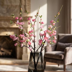 May include: A tall, dark gray glass vase holds several branches of artificial pink cherry blossoms. The branches feature textured brown stems, small green leaves, and numerous delicate pink flowers in various stages of bloom, from tight buds to fully open petals.