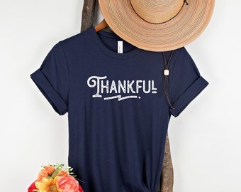 May include: A navy blue t-shirt with the word "THANKFUL" printed in white on the front. The shirt is hanging on a wooden ladder with a straw hat on top.