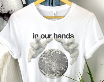 May include: White t-shirt with a graphic of a globe surrounded by two hands, and the text "in our hands".
