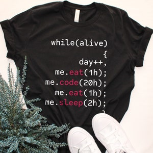 Könnte beinhalten: Schwarzes T-Shirt mit weißem Text, der "while(alive) { day++; me.eat(1h); me.code(20h); me.eat(1h); me.sleep(2h); }" in einer code-ähnlichen Schriftart lautet. Der Text ist pink.