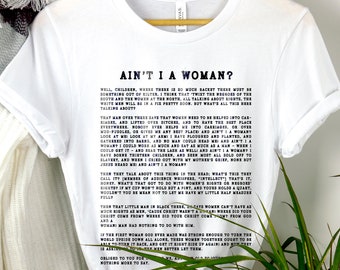 以下が含まれることがあります： 黒い文字で「Ain't I a Woman?」と書かれた白いTシャツ。このテキストは、1851年にオハイオ州アクロンの女性大会でソジャーナー・トゥースが演説したものです。この演説は、女性の権利と平等についてです。