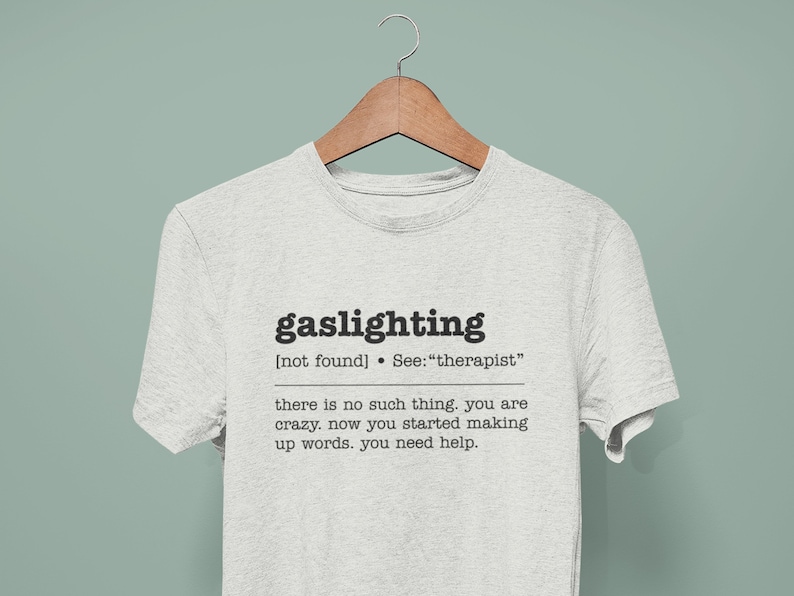 以下が含まれることがあります： 「ガスライティング」という言葉が定義されたグレーのTシャツ。「[見つかりません] 参照: 「セラピスト」」と「そんなものはありません。あなたは狂っています。あなたは今、言葉をでっち上げ始めました。あなたは助けが必要です。」というテキストが書かれています。
