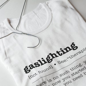 以下が含まれることがあります： 「ガスライティング」という言葉が定義された白いTシャツ。「[見つかりません]。参照：「セラピスト」そんなものはありません。あなたは今、作り始めています。助けが必要です。 」
