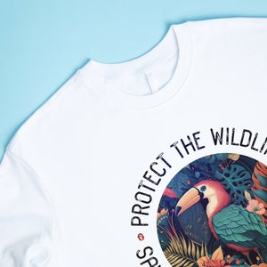 May include: White t-shirt with a circular graphic design featuring a toucan in a jungle setting. The text "PROTECT THE WILDLIFE" is printed around the circle.