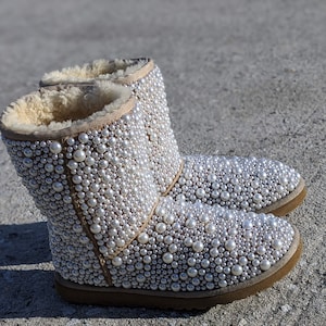 May include: A pair of beige boots covered in faux pearls. The boots have a light brown sole and a tan interior. The exterior is completely covered in various sizes of white pearls, creating a textured, glamorous look.