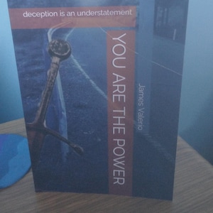 Può includere: Una copertina di libro con sfondo blu e una strada sullo sfondo. Il testo sulla copertina recita "deception is an understatement" e "You Are The Power" con il nome dell'autore "James Valerio" sotto.