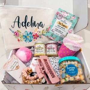May include: A pink and white gift box filled with various items, including a personalized canvas bag with the name "Adelina" embroidered on it, a face mask, a body scrub, a bath bomb, a hair clip, a scrunchie, a cupcake-shaped container of candy, and a small card with the text "A little something to brighten your day".