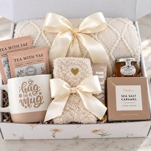 May include: A gift box containing a cream-colored textured throw blanket with a satin ribbon, two Tea With Tae packets, a speckled ceramic mug with the text Hug in a Mug, fuzzy socks with a gold heart, a jar of honey, and Sea Salt Caramel cookies.