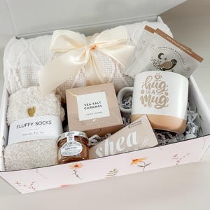 May include: A gift box filled with a white blanket, fluffy socks, a mug with "hug in a mug" text, tea, sea salt caramel cookies, honey, and shea butter. The box is decorated with floral patterns and a cream-colored bow.