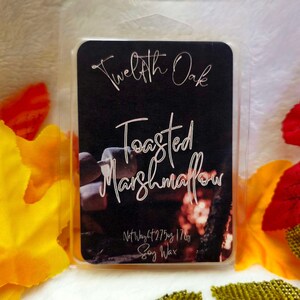 May include: A black wax melt packaged in clear plastic, labeled "Twelfth Oak Toasted Marshmallow Soy Wax." The label features white script and a blurred image. The background includes colorful fall leaves.