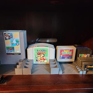 May include: A collection of video game cartridges organized in a gray plastic storage unit. The cartridges are for the Nintendo 64 and Super Nintendo Entertainment Systems. The cartridges include "Banjo-Tooie", "Mario's Tennis", and "Golden Sun".