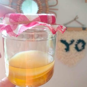 Pode incluir: Um frasco de vidro com uma tampa de tecido xadrez vermelho e branco, cheio de um líquido amarelo claro e um objeto redondo branco flutuando. O frasco está sendo segurado por uma mão.