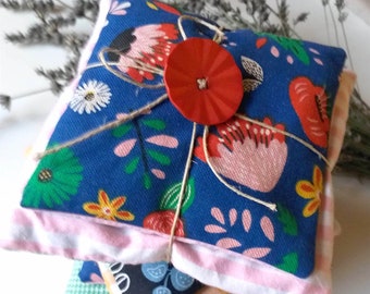 Puede incluir: Un conjunto de cuatro cuadrados de tela con un patrón floral. El cuadrado superior está atado con una cuerda y tiene un botón rojo en el centro. Los cuadrados son en tonos de azul, rosa, verde y naranja.