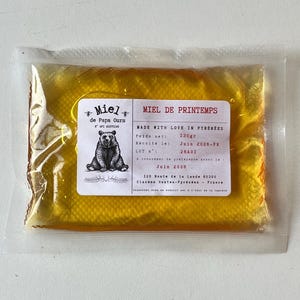 Peut inclure: Un sachet rectangulaire en plastique transparent contenant du miel jaune doré. Une étiquette blanche présente une illustration d'ours noir et le texte Miel de Papa Ours, Miel de Printemps, Made with Love in Pyrenees, 130gr, et Juin 2028.