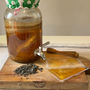 Peut inclure: Un grand distributeur de boissons en verre rempli de liquide ambré et d'une mère de kombucha, surmonté d'une housse en tissu à motifs verts et blancs. À côté se trouvent du thé en vrac, deux sachets en plastique scellés et une cuillère à miel.