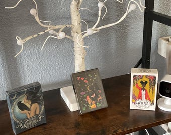 May include: Three different tarot card decks on a wooden shelf. The decks are titled "Mystic Sisters", "Moon Witch", and "Witch of the Wildwood". A white birch tree with white lights is on the shelf behind the decks.