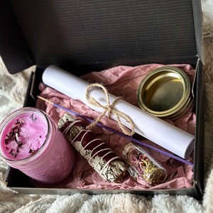 May include: A gift box filled with a pink candle, a white sage smudge stick, a small glass jar of dried herbs, a rolled up piece of paper tied with twine, and a gold lid.