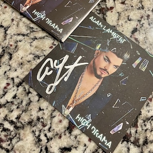 May include: A signed CD by Adam Lambert, titled "High Drama". The CD cover features a black and white photo of Adam Lambert with a silver chain necklace. The background is a dark, abstract design with iridescent accents.