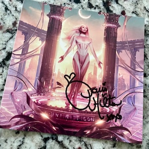 May include: A signed photograph of a woman in a white outfit standing on a platform with a futuristic cityscape in the background. The signature is "Paris Hilton" with "XP" below it. The text "INFINITE ICON" is visible at the bottom of the image.
