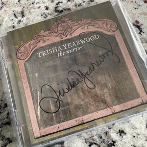 May include: A CD case featuring the album "the mirror" by Trisha Yearwood. The album cover has a vintage aesthetic with a pink-framed mirror design and the artist's signature. The CD is sealed in clear plastic.
