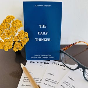 May include: A navy blue 2026 desk calendar titled "The Daily Thinker" with carefully curated quotes. The image includes a pen, eyeglasses, and cards with quotes from famous thinkers. A bouquet of yellow flowers is also present.