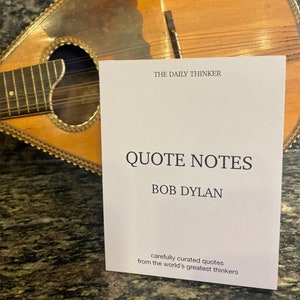 May include: A white card with the text "THE DAILY THINKER" and "QUOTE NOTES" in black font. Below the title is the text "BOB DYLAN" in black font. The card also includes the text "carefully curated quotes from the world's greatest thinkers". The card is sitting on top of a wooden mandolin.