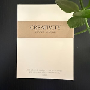 May include: A stack of cream-colored quote notes with a brown band that reads "CREATIVITY QUOTE NOTES." The bottom of the notes has the quote "Art should comfort the disturbed and disturb the comfortable." by Banksy.