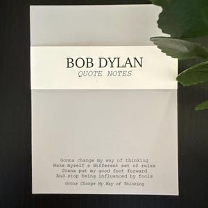 May include: A white notepad with the text "BOB DYLAN QUOTE NOTES" printed in black. The notepad features a quote by Bob Dylan. The quote is printed in a typewriter-style font. The notepad is on a black surface.