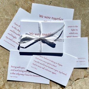 May include: A stack of white cards with red ink, tied together with a white ribbon. Each card features a different quote about love and togetherness. The quotes include: "We were together", "yours is the light by which my spirit's born", "Being with you and not being with you is the only way I have to measure", "Or never before in story or rhyme (Not even once upon a time) Has the world ever known a you, my friend, And it never will, not ever again."