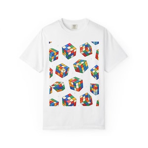 May include: White t-shirt featuring a colorful pattern of Rubik's Cubes. The cubes are rendered in a variety of orientations and feature the classic color scheme of red, blue, green, yellow, orange, and white. The shirt is a casual, short-sleeved style.