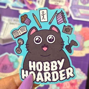 Könnte beinhalten: Ein Aufkleber mit einer Cartoon-Katze und dem Text "HOBBY HOARDER". Die Katze ist von Illustrationen von Hobbys wie einem Cupcake, einer Kamera, einer Hantel und einem Gamecontroller umgeben. Der Aufkleber hat einen blauen Hintergrund.
